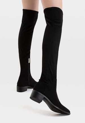 Benen met zwarte over-de-knie laarzen met lage blokhakken en zijritsen, afgebeeld tegen een effen lichte achtergrond.