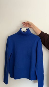 Blauwe coltrui van gebreide stof, met een geribde kraag en lange mouwen, getoond op een witte hanger tegen een effen achtergrond.