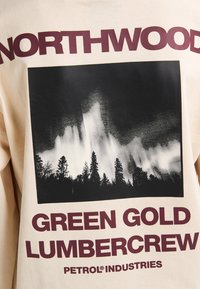 Felpa beige con una grafica in bianco e nero di alberi sotto un cielo stilizzato. Il testo recita "NORTHWOOD" e "GREEN GOLD LUMBERCREW."