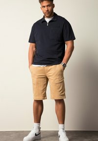 Polo bleu marine à manches courtes avec un col zippé et une poche, associé à un short cargo beige. Le modèle porte des baskets blanches et des chaussettes. Fond simple.