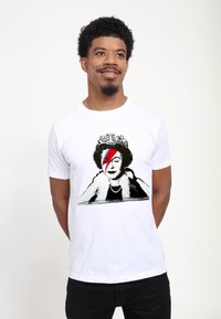 Bílý bavln�ěný tričko s grafikou ženy s korunou a designem červeného a modrého blesku, připomínající pop art.