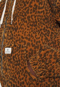 Ruskea corduroy-ziippihuppari, jossa on musta leopardikuvio. Hupparissa on etutasku, valkoiset nyörit ja ribbaus hihansuissa.