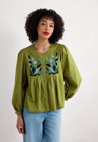 Zelená bavlněná blůza s nab íranými rukávy, zapínáním na knoflíky vpředu a květinovou výšivkou modré a tmavě zelené barvy na hrudi. Nosená s světle modrými džíny.