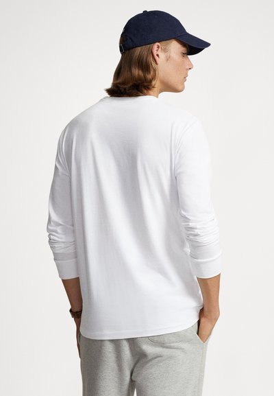 Ilgas rankovėmis baltas medvilninis marškinėlis klasikiniu siluetu, su apvaliu iškirpimu ir rankogaliais, derinamas su šviesiai pilkomis sportinėmis kelnėmis ir tamsiai mėlyna kepure.