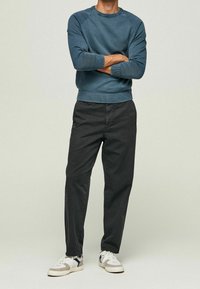 Muž stojící s rukama zkříženýma, oblečený v modrém svetru s dlouhým rukávem, černých kalhotách a bílých teniskách na jednobarevném pozadí.