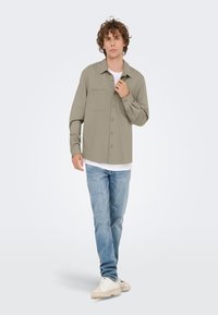 Lichtgrijs overhemd met knopen, een borstzak, lange mouwen en een losse pasvorm, gecombineerd met lichtblauwe jeans en beige sneakers.