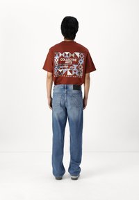 Brown short-sleeve t-shirt with a geometric patterned graphic on the back, paired with loose-fitting blue jeans and black shoes.