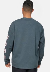 Pitkähihainen paita haalistuneessa teal-värissä, jossa on pyöreä kaula-aukko ja painettu teksti vasemmassa hihassa. Valmistettu pehmeästä, teksturoidusta puuvillamateriaalista.