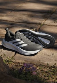 Chaussures de course Adidas grises et blanches avec semelles texturées reposant sur un chemin en pierre ensoleillé avec de petites plantes à proximité.