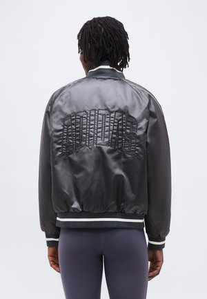 Isik tume punutud juustega, kandes musta Adidas siidist bomberjopet, mille seljal on tikitud logo, kombineeritud tumedate retuusidega.