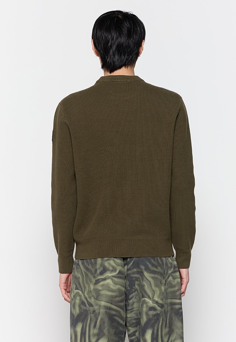 Maglione a coste verde oliva con scollatura rotonda e maniche lunghe. Indossato con pantaloni larghi e a fantasia. Nessun hardware o accento visibile.