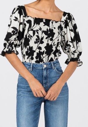 Femme portant un haut à manches bouffantes noir et blanc à motif floral avec corsage smocké et jean bleu taille haute, mains jointes à la taille.