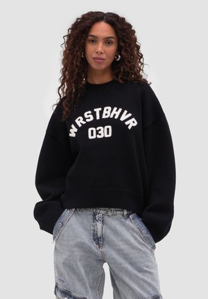 Frau mit lockigem Haar, die einen schwarzen Pullover mit dem Schriftzug "WRSTBHVR 030" und hellblaue weite Jeans trägt, steht mit den Händen in den Taschen.