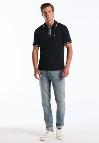 Marineblauwe polo met witte accenten, korte mouwen en een voorzak, gecombineerd met lichtblauwe jeans en witte sneakers. Effen achtergrond.