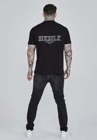 Črna kratkotrajna majica s kratkimi rokavi z odtisnjenim logotipom "SIKSILK" na hrbtni strani. Ujemajoč s črnimi kavbojkami in belimi supergami. Gladka tekstura, sproščen kroj.