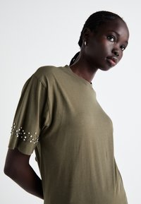 Olijfgroene T-shirt met een relaxed fit, korte mouwen en decoratieve juwelenaccenten op de mouw, gemaakt van zacht materiaal.