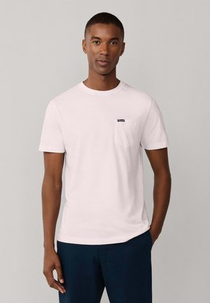 Ein Mann steht frontal mit einem schlichten weißen Kurzarm-T-Shirt mit einer kleinen Brusttasche und dunkelblauen Hosen vor einem grauen Hintergrund.