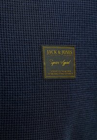 Tissu texturé bleu foncé avec une étiquette noire et dorée Jack & Jones indiquant "Superior Apparel" et des détails de design en petits caractères.
