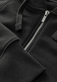 Čierna mikina na zips vyrobená z mäkkého materiálu. Obsahuje rebrované manžety a šnúrku kapucne s textúrou. Strieborný metalový zips pozdĺž prednej strany.