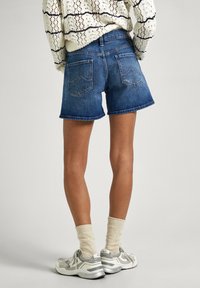 Jeans kratke hlače v srednji modri barvi z raztrganim robom, z dvema zadnjima žepoma s ukrivljenim šivom. Nosijo se z bež nogavicami in srebrnimi supergami.