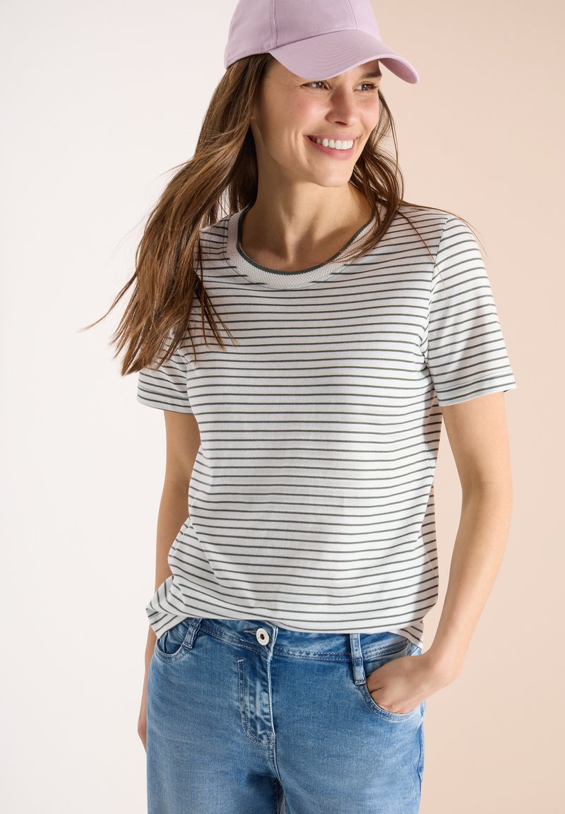 Femme souriante portant une casquette rose, un t-shirt blanc à rayures, et un jean bleu, une main dans la poche, regardant sur le côté.