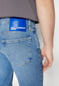 Blå denimjeans har en synlig bakficka och en blå läderlapp märkt "KARL LAGERFELD JEANS." Ljust tvättad med synlig sömnad.