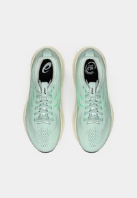 Ténis atléticos em azul claro com parte superior em rede, destaques verdes e sola almofadada. Os atacadores são cinzentos; as palmilhas apresentam um logótipo.