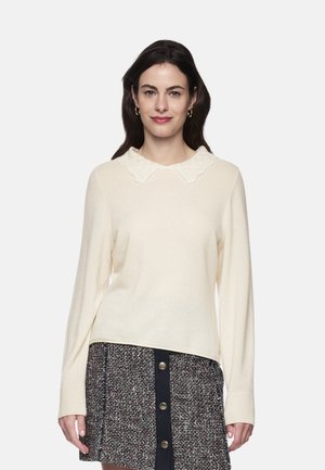 Woman with dark hair wears a cream sweater with a textured collar and a buttoned tweed skirt against a plain light background.