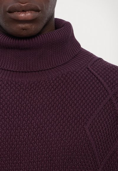 Pull à col roulé violet foncé avec un motif de tricot texturé et un col côtelé épais. Présente des coutures inclinées sur les épaules.