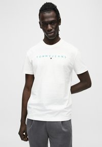 Λευκό βαμβακερό μπλουζάκι με στρογγυλή λαιμόκοψη και κοντά μανίκια, με την επιγραφή "TOMMYJEANS" σε τυρκουάζ χρώμα και ένα μικρό λογότυπο σε κόκκινο, λευκό και μπλε.