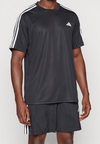 Černé sportovní tričko s bílými pruhy na ramenech, vyrobené z tkaniny odvádějící vlhkost. V páru s odpovídajícími černými šortkami se zvýrazněním loga.