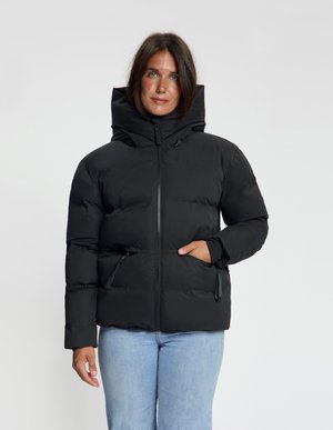 Vrouw draagt een zwart donsjack met een hoge kraag en rits, staat met één hand in de zak en lichtblauwe spijkerbroek.