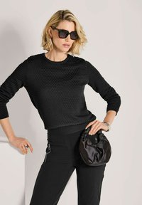Frau mit kurzen blonden Haaren, die einen schwarzen strukturierten Pullover, schwarze Hose mit Reißverschlussdetails, schwarze Sonnenbrille trägt und eine schwarze Satinhandtasche hält.