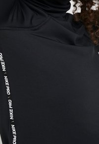 Schwarzes Nike Pro Langarmshirt aus glattem, dehnbarem Material. Verfügt über einen Stehkragen und einen weißen Logostreifen an der linken Seite.