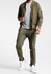 Olivově zelená bomber bunda, světlé šedé tričko s malými černými vzory, olivové kalhoty a bílé tenisky. Texturovaná tkanina se zipy.