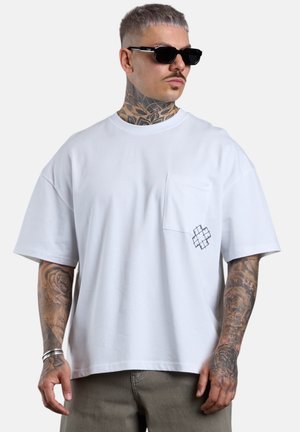 Mann mit Tätowierungen und Sonnenbrille trägt ein weißes Oversize-T-Shirt mit Brusttasche und kleinem schwarzem Kreuz-Design.