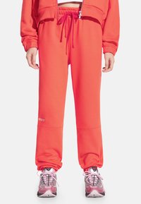 Jasnoróżowe spodnie dresowe w kolorze koralowym z elastycznym pasem i sznurkiem, wyposażone w prążkowane mankiety oraz logo przy krawędzi. Połączone z różowymi sneakersami.