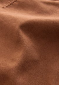 Brauner Wildlederstoff mit einer weichen, strukturierten Oberfläche, die sanfte Falten und Knicke zeigt und das Licht subtil über das Material reflektiert.