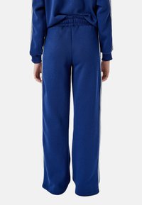 Blaue Jogginghose mit lockerer Passform, elastischem Bund und weißen Seitestreifen. Hergestellt aus weichem, strukturiertem Stoff. Keine sichtbaren Taschen.