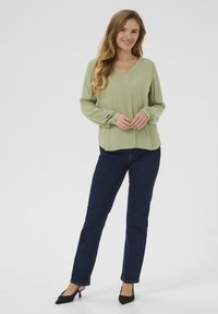 Blouse vert clair à manches longues avec un col en V et des poignets boutonnés, associée à un jean ajusté bleu foncé et des talons noirs.