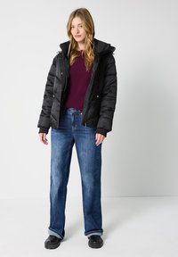 Sort pufferjakke med hætte, lynlås på fronten og elastiske mansjetter. Bæres over en bordeaux sweater, parret med løse blå jeans og sorte sko.