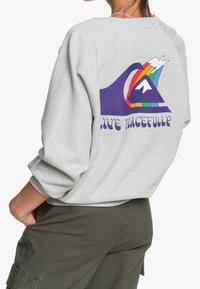 Sudadera gris con un gráfico de olas coloridas y el texto "VIVE EN PAZ" en púrpura. Hecha de forro polar, con un corte relajado y puños acanalados.