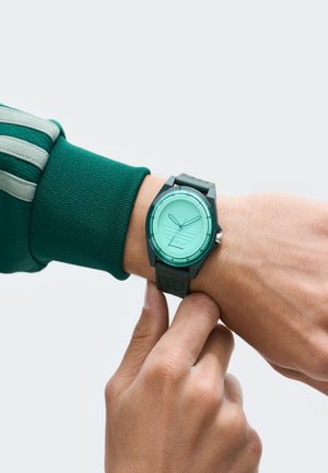 Cilvēks ar zaļo garo piedurknēm uzvalku pielāgo melno un zaļo rokas pulksteni ar tirkīza krāsas ciparnīcu, kas rāda laiku.