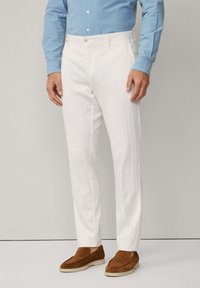 Pantalons blancs ajustés avec une texture lisse, dotés d'une fermeture à bouton et de poches latérales, associés à des chaussures à enfiler en daim marron.