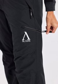Schwarze Cargo-Hose aus glattem Stoff, mit einer Reißverschlusstasche, schrägen Nähten und einem auffälligen weißen "SLADE"-Logo.