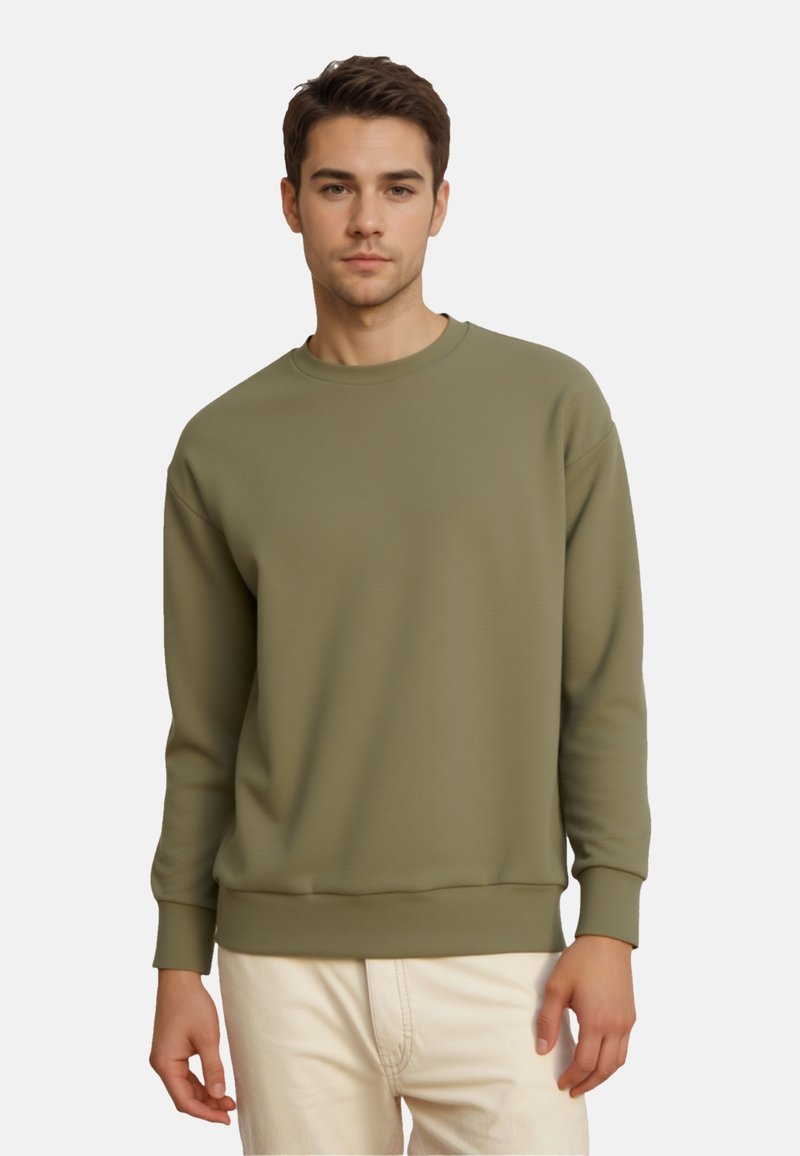 Jeune homme portant un sweat-shirt vert olive à col rond avec des manches longues et un pantalon beige clair, debout devant un fond uni.