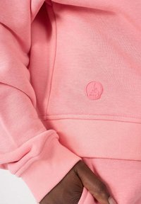 Felpa rosa chiaro con una texture morbida, polsini a coste e un piccolo patch logo rosa nella zona della tasca frontale.