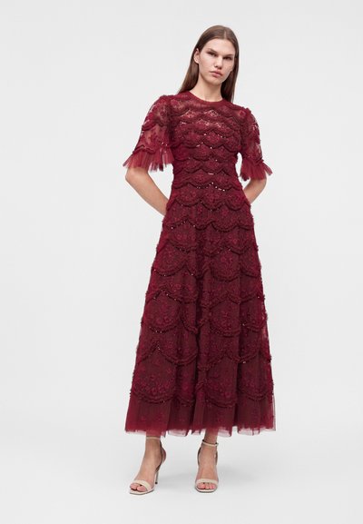 Femme portant une robe en dentelle bordeaux avec des motifs floraux festonnés et des manches courtes transparentes, associée à des talons ouverts beiges, debout sur un fond blanc.