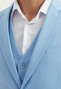 Veste de costume bleu clair avec revers crantés, deux boutons et un mouchoir de poche. Chemise habillée blanche en dessous avec une texture unie.