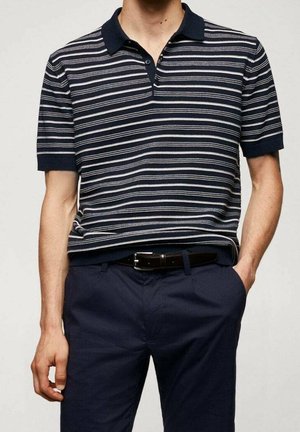 Mężczyzna ubrany w granatowo-biały paskowany, krótki rękaw polo, włożony do ciemnogranatowych spodni z czarnym paskiem, z jedną ręką w kieszeni.